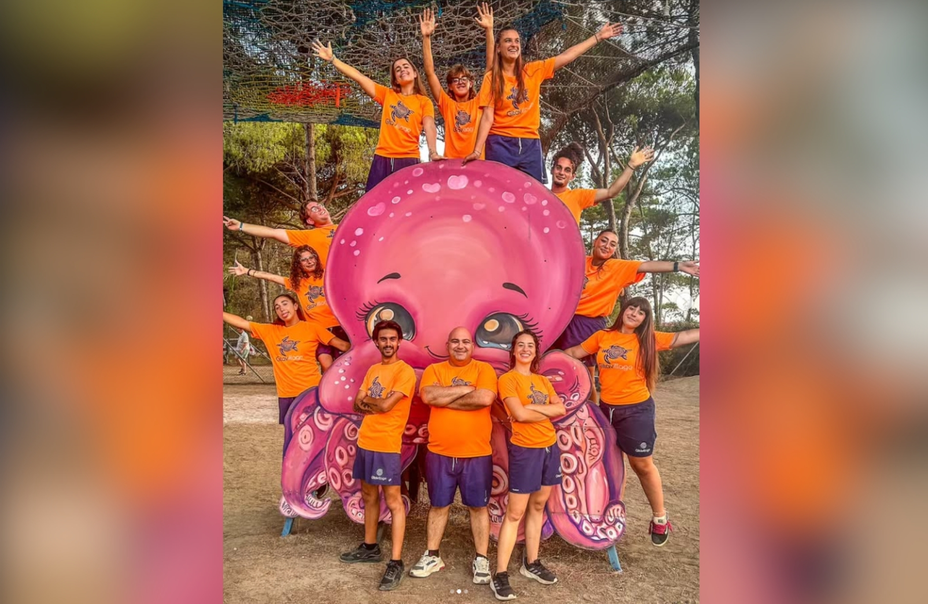 Un gruppo di persone in magliette arancioni posano accanto a una grande statua di polpo rosa in un parco, sorridono e alzano le braccia.