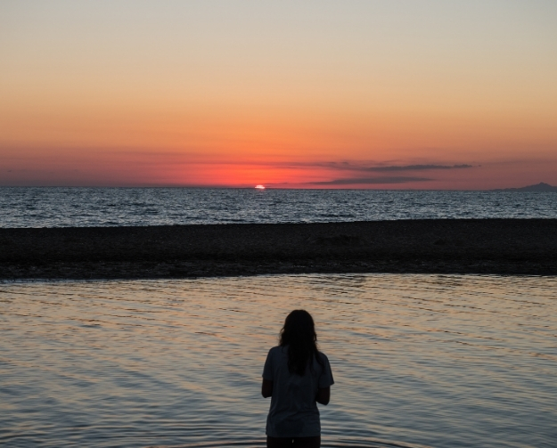 Una persona di spalle è in acqua, davanti al mare, mentre il tramonto tinge il cielo di arancione e rosa.